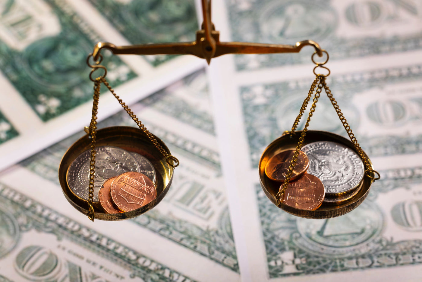 Close-up of a vintage balance scale with coins of different values on each side, set against a background of U.S. dollar bills, symbolizing financial balance, inequality, or investment decisions.