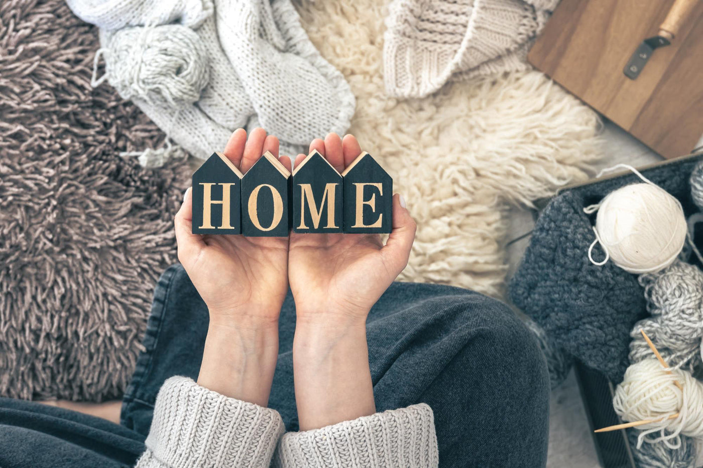 Close-up of hands holding wooden house-shaped blocks spelling 'HOME,' surrounded by cozy knitted blankets and yarn in a warm, inviting setting.