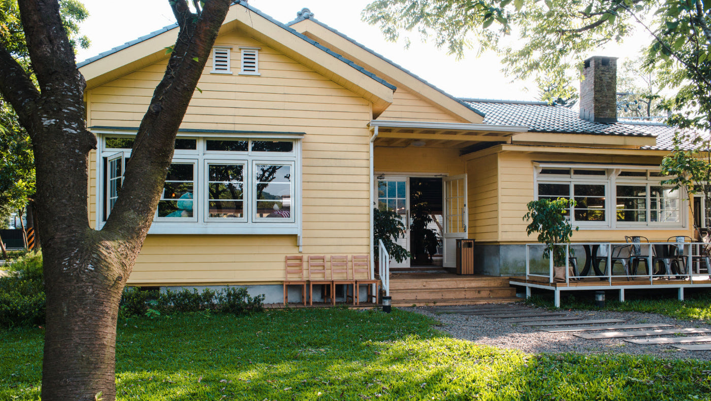 A charming yellow single-story house with white trim, a front porch, and a lush green lawn, surrounded by trees and outdoor seating.