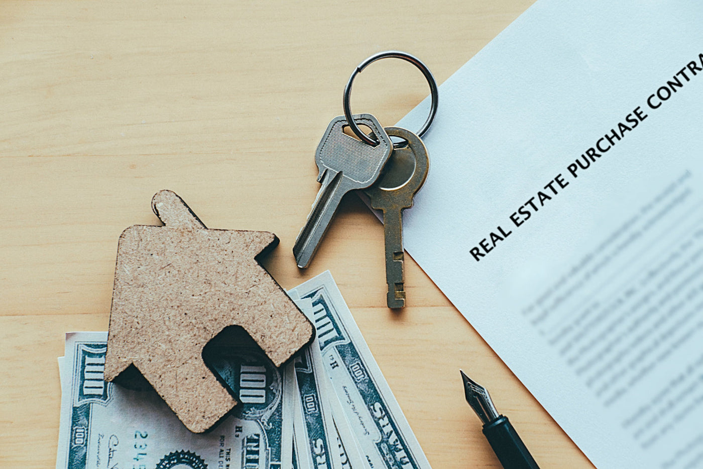 A real estate purchase contract on a wooden desk, accompanied by a set of house keys, a small wooden house-shaped model, cash, and a fountain pen, symbolizing a home buying process.