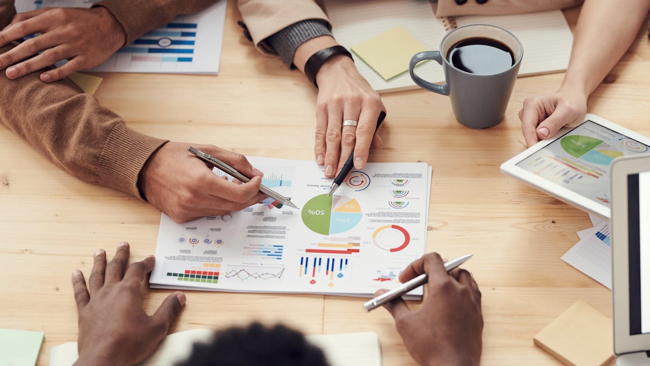 This image shows a collaborative business meeting with people analyzing charts and graphs, discussing data insights, and using digital devices.