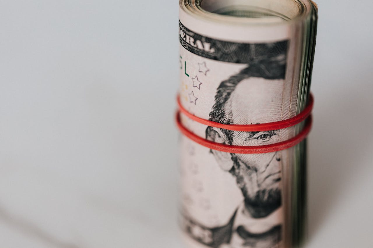 A rolled stack of U.S. five-dollar bills secured with a red rubber band, placed on a white surface.