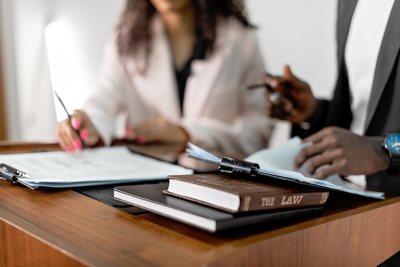 Two professionals engaged in a discussion over legal documents at a wooden desk, with a book titled 'The Law' and a clipboard with papers in the foreground, symbolizing a legal consultation or business agreement.