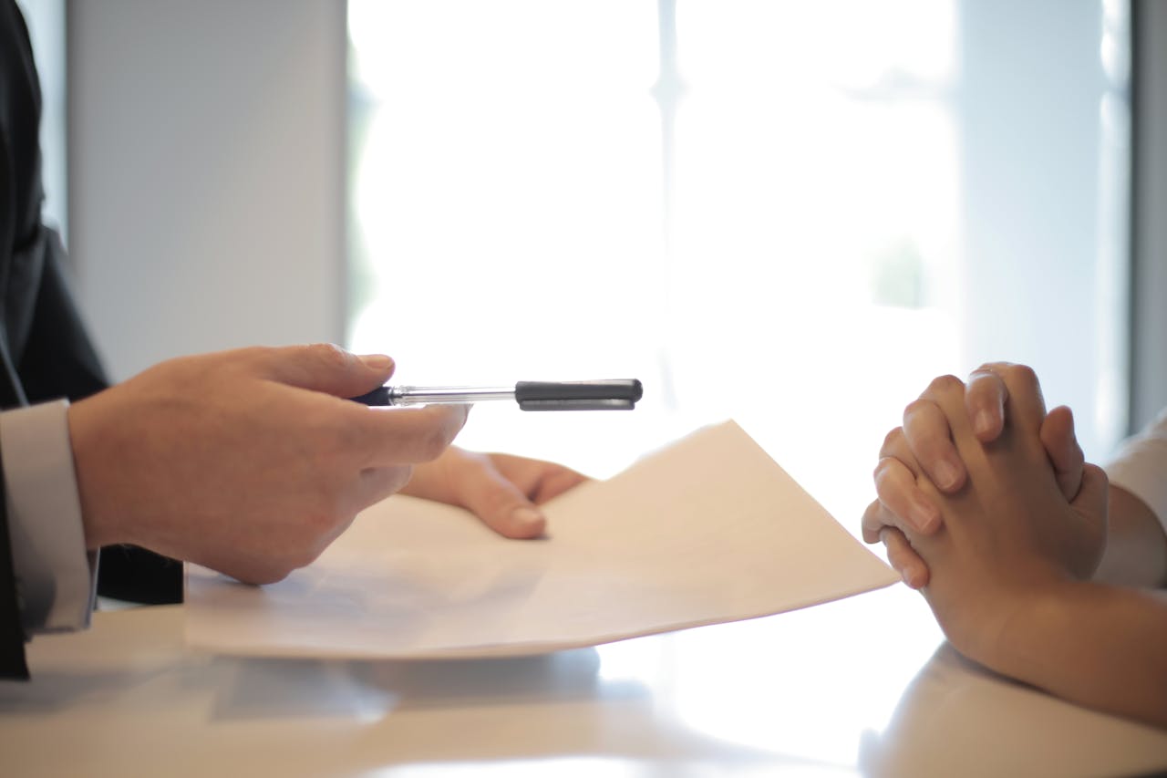 Close-up of a businessperson handing a pen and contract to another person with clasped hands, suggesting a signing or negotiation process.