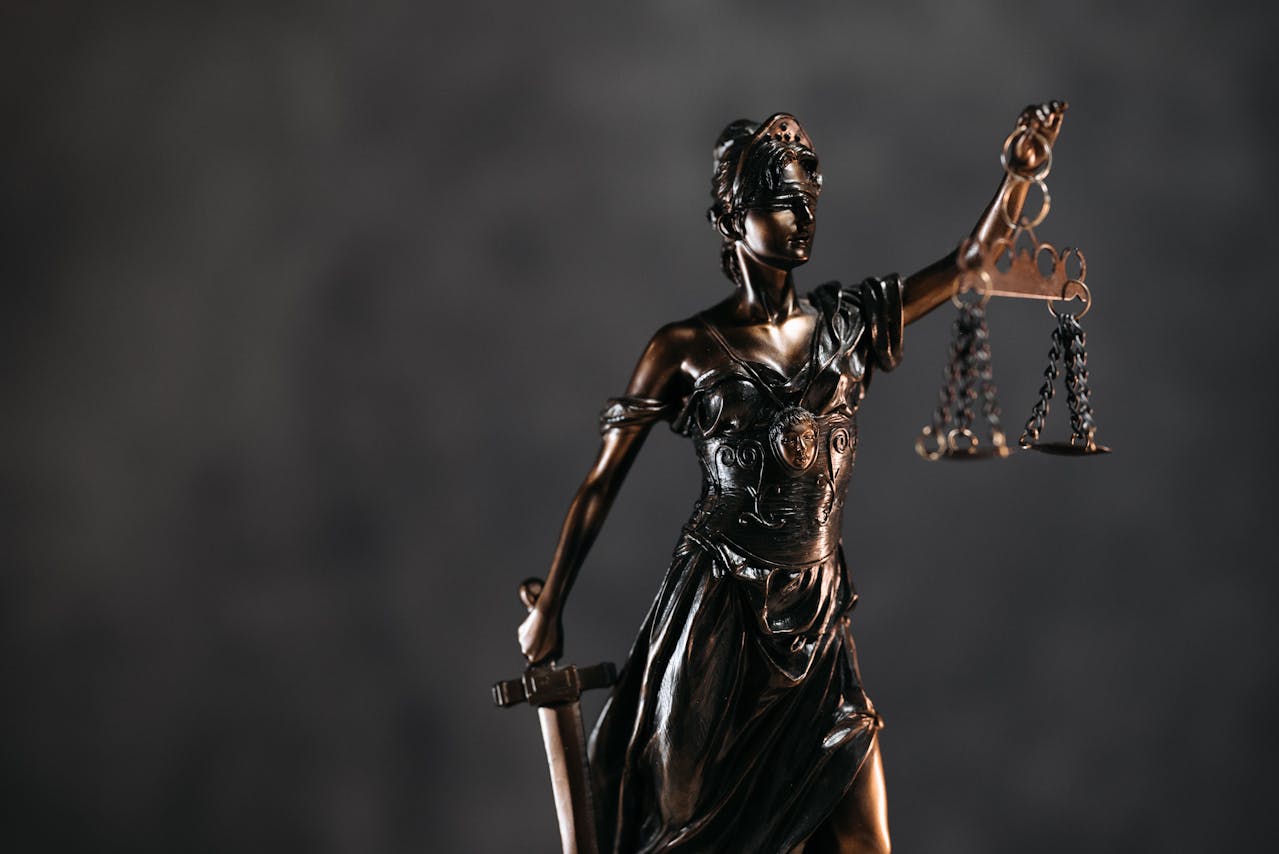 A close-up of a bronze statue of Lady Justice, blindfolded and holding a set of scales in one hand and a sword in the other, symbolizing fairness and the rule of law, against a dark, blurred background.