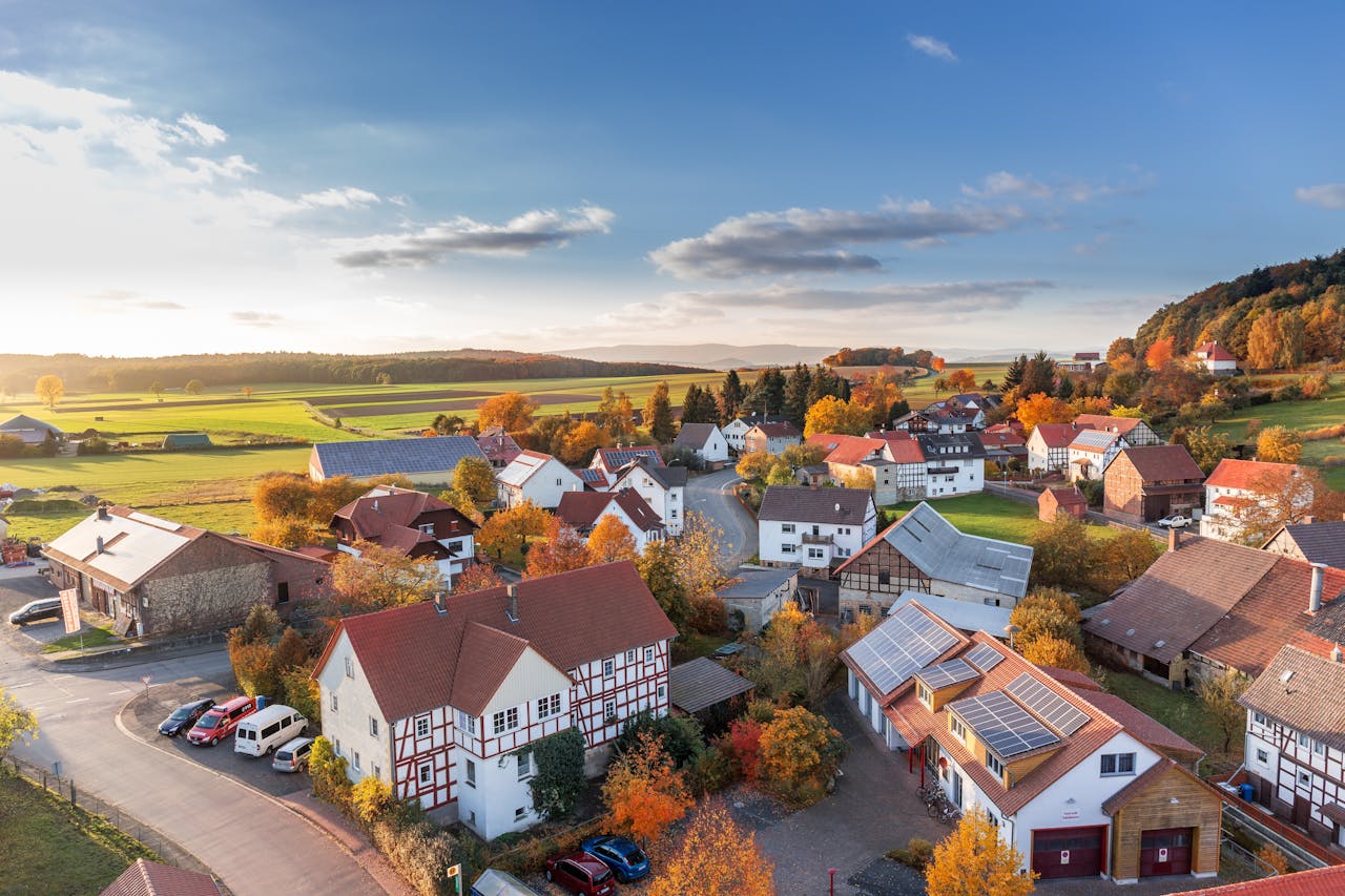 Aerial view of a picturesque village surrounded by rolling green fields and autumn-colored trees, featuring traditional houses with red roofs, solar panels, and winding roads under a bright blue sky.