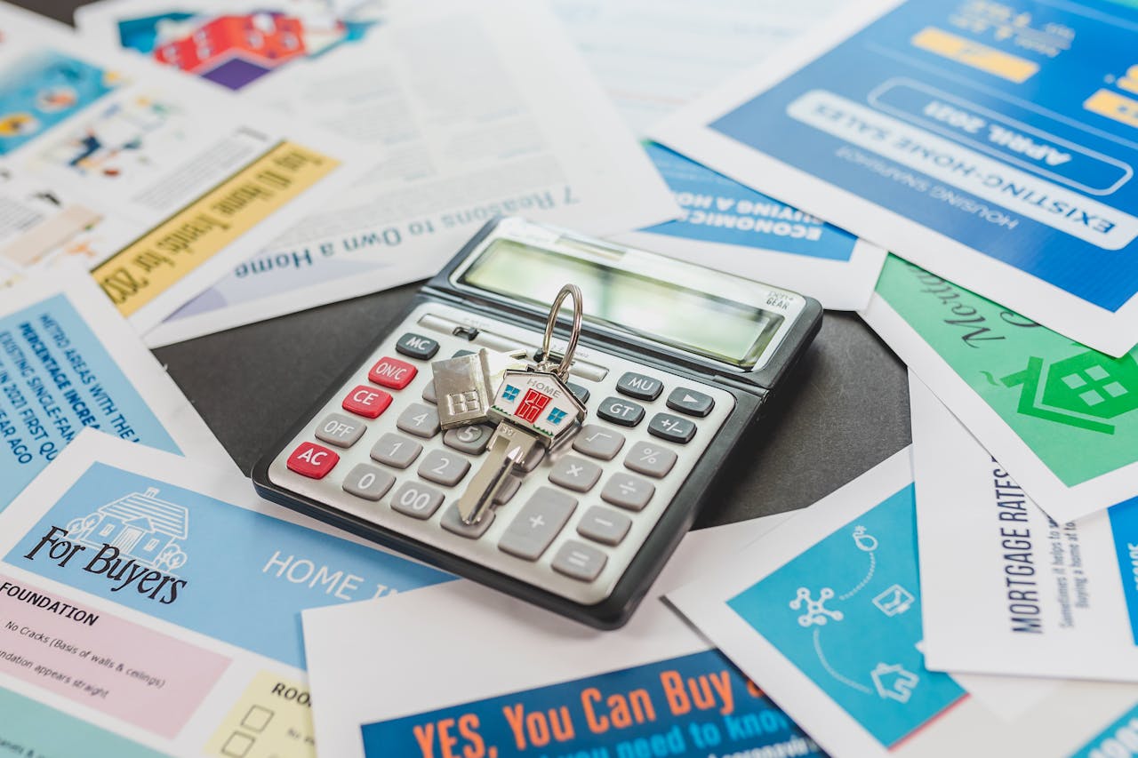 A calculator with a house-shaped keychain on top, surrounded by real estate documents, mortgage information, and buyer guides, representing financial planning, home buying, or real estate investment.