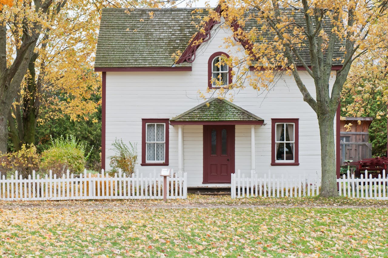 A charming white cottage with red trim, a mossy roof, and a small porch, surrounded by a white picket fence.